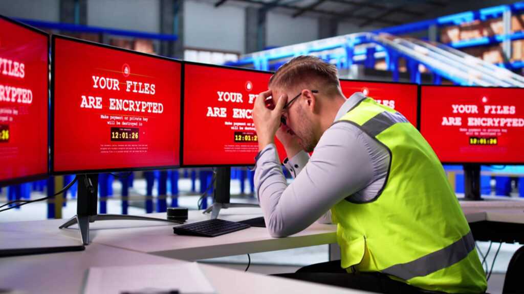 Frustrated employee in office facing multiple monitors displaying red ransomware encryption warnings representing cyber attacks